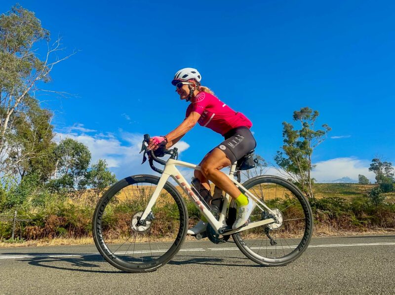 A woman in a pink cycling jersey, black shorts, and a white helmet is riding a white bicycle on a paved road under a blue sky. The road is surrounded by dry grasses and trees, with rolling hills in the background.