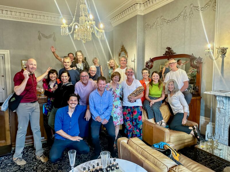 Final dinner group photo of guests in casual attire smiling and posing inside a historic room at Lenna of Hobart, which features chandeliers, a large mirror, and ornate wallpaper. The Strzelecki Message Stick is displayed by one of the guides