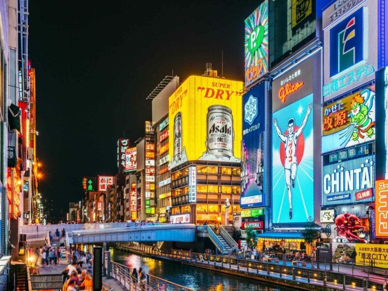 A city street with tall, brightly lit buildings covered in billboards, with a canal in place of a road up the middle. A footbridge joins the two sides of the street. People can be seen walking along the street and bridge.