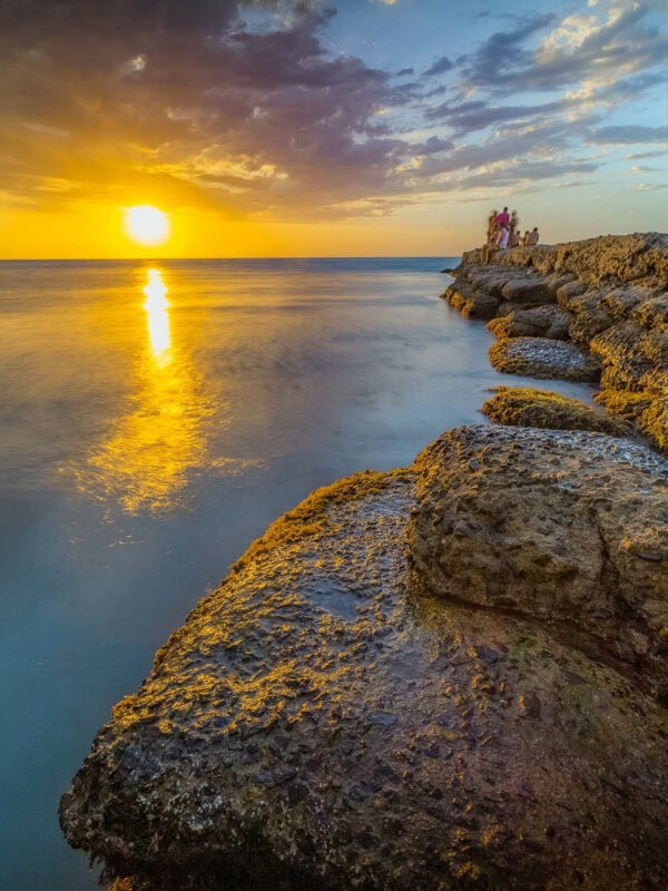 Golden Caribbean sunset reflecting on calm ocean waters with people on coastal rocks.
