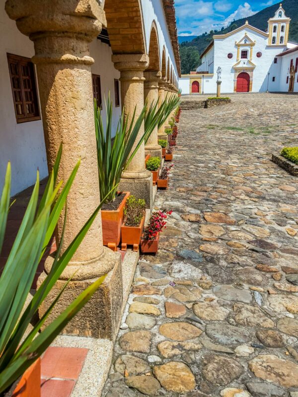 Stone courtyard lined with colonial arches and potted plants, leading toward a white church with red doors.