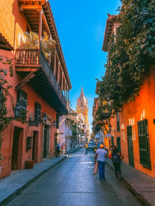 Narrow street in Cartagena with colonial balconies and colourful facades, ending with the cathedral tower in the distance.