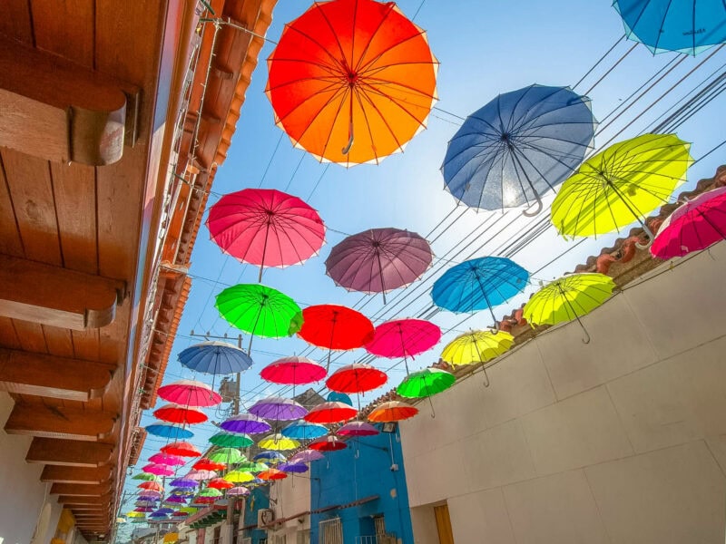 Colourful umbrellas hanging above a narrow street, creating a playful canopy under bright blue skies.
