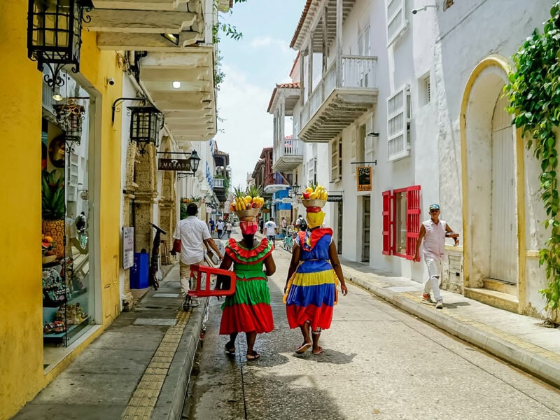 Two Palenqueras in traditional dresses carrying bowls of fruit on their heads while walking through Cartagena’s old town.