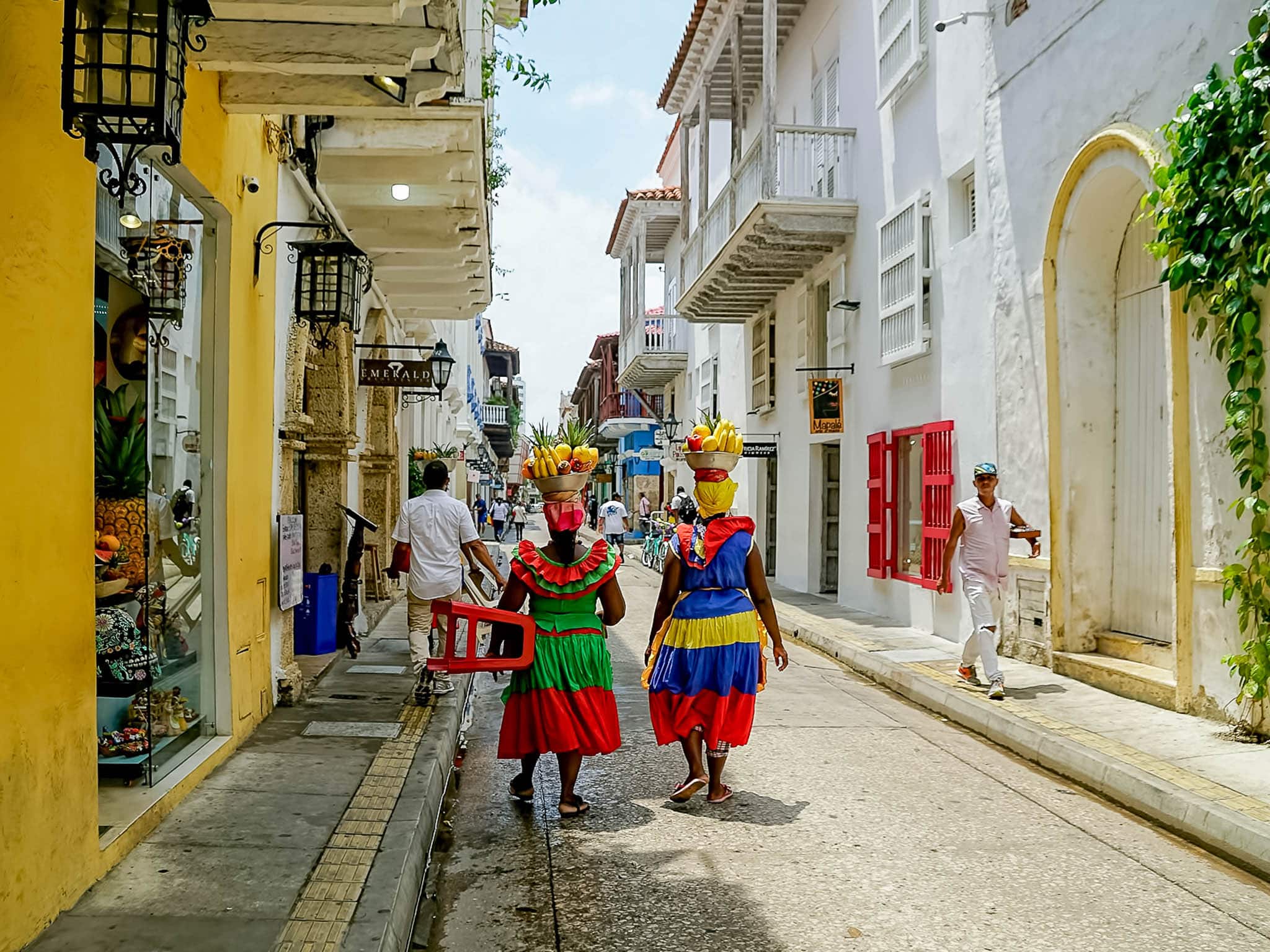 Two Palenqueras in traditional dresses carrying bowls of fruit on their heads while walking through Cartagena’s old town.