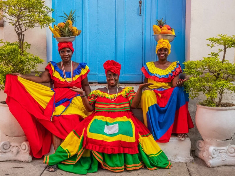 Three women in vibrant traditional dresses, two with bowls of fruit on their head dresses, smiling in front of a bright blue doorway.