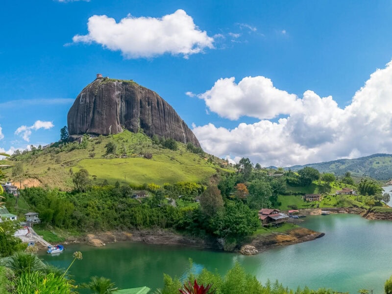 The Rock of Guatapé (El Peñol) rising tall above the green hills and lake below on a clear day. There are houses dotted along the shore of the lake.