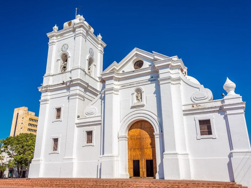 Whitewashed Santa Marta Cathedral with twin towers and a large wooden entrance door against a deep blue sky.
