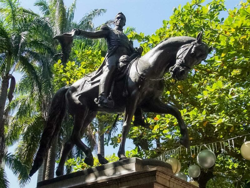 Equestrian statue of Simón Bolívar in military attire of the day, holding out his hat. The statue is surrounded by palm and other trees in a sunlit plaza.
