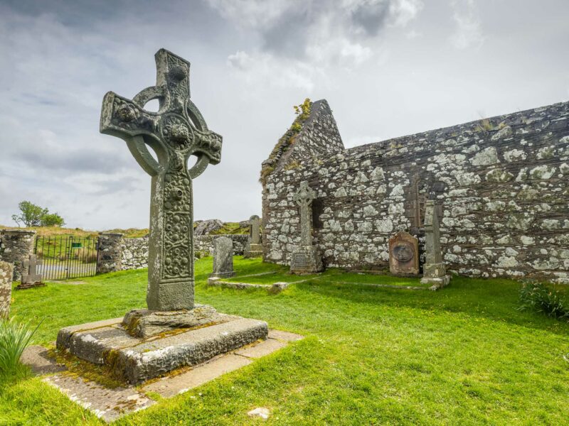 Ancient carved Celtic cross standing near ruined stone walls of an old abbey on grassy ground.