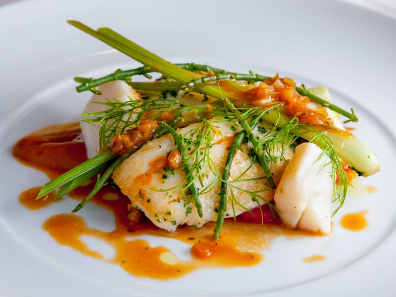 Seared fish fillet with asparagus spears, herbs, and a colourful sauce, elegantly presented on a white plate.