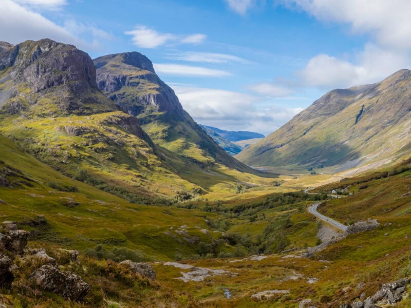 Wide glen with winding road, dramatic mountain peaks, and golden sunlight breaking through clouds.