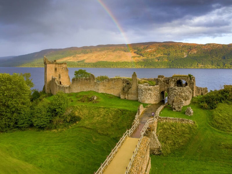 Medieval castle ruins perched on a cliff above the river, with a rainbow arching across the sky and a low ridge of hills in the background.