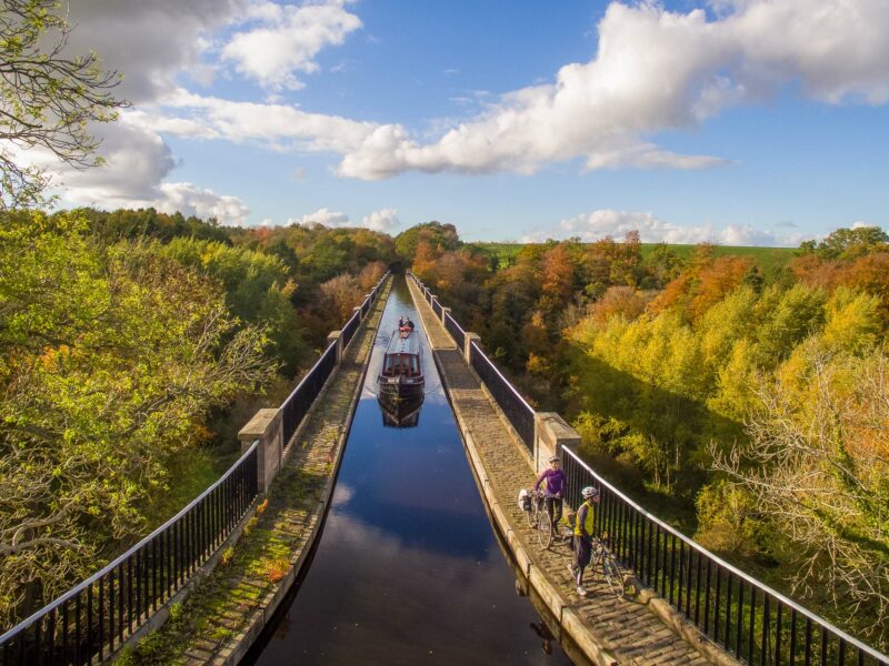 Narrow canal boat gliding across a tall stone aqueduct surrounded by autumn-coloured trees.