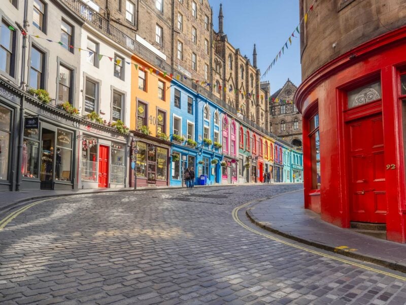 Row of colourful Georgian shopfronts and cafés on a curved cobblestone street in Edinburgh’s Old Town.