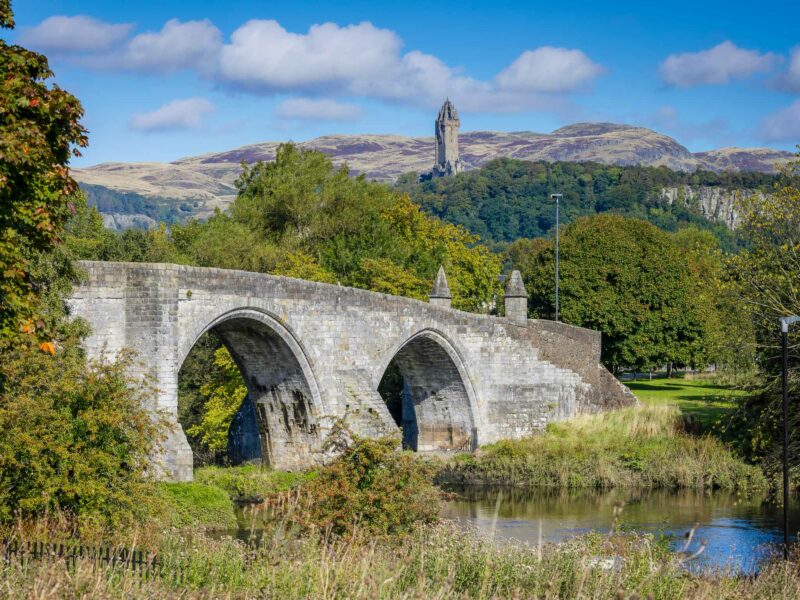 Old arched stone bridge crossing a calm river, with forested hills behind and bare rocky mountains in the distance.