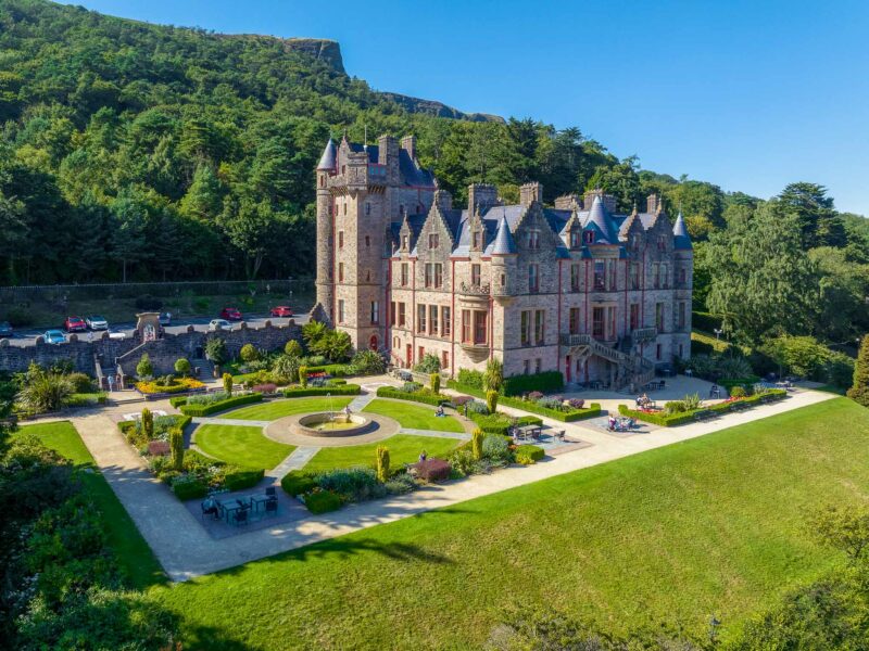 Large baronial castle with towers, turrets, and landscaped gardens, set against a backdrop of wooded hills and a deep blue sky