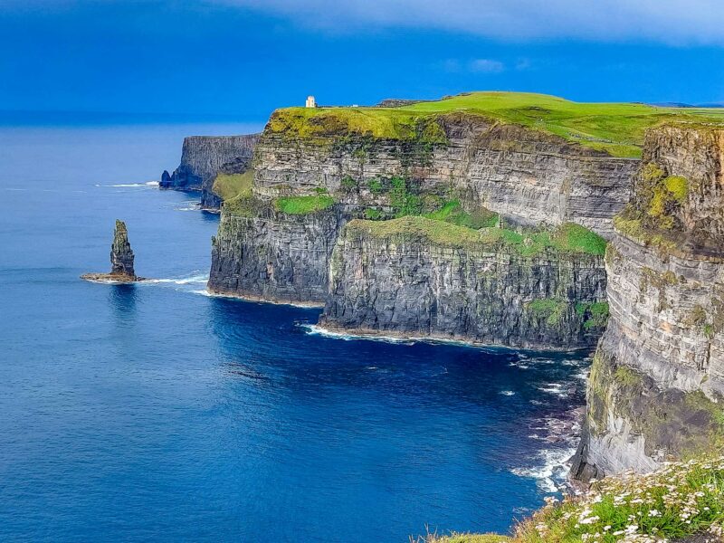 Cliffs rising dramatically over the Atlantic Ocean with waves crashing below. The deep blue ocean meets the vivid grey blue skies on the horizon in the background