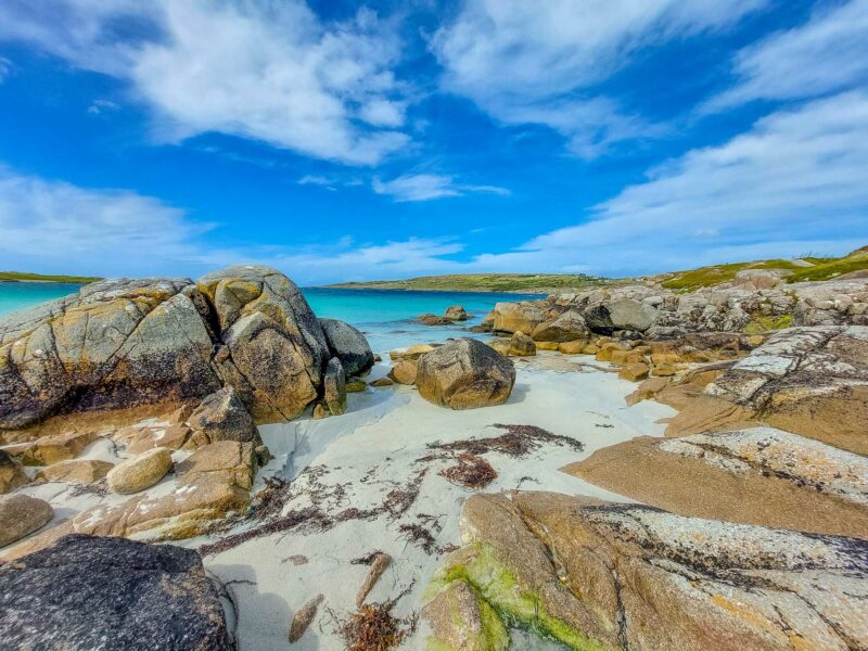 Jagged rocks sit large among turquoise tidal pools and sandy stretches, framed by distant hills under a wide open sky.