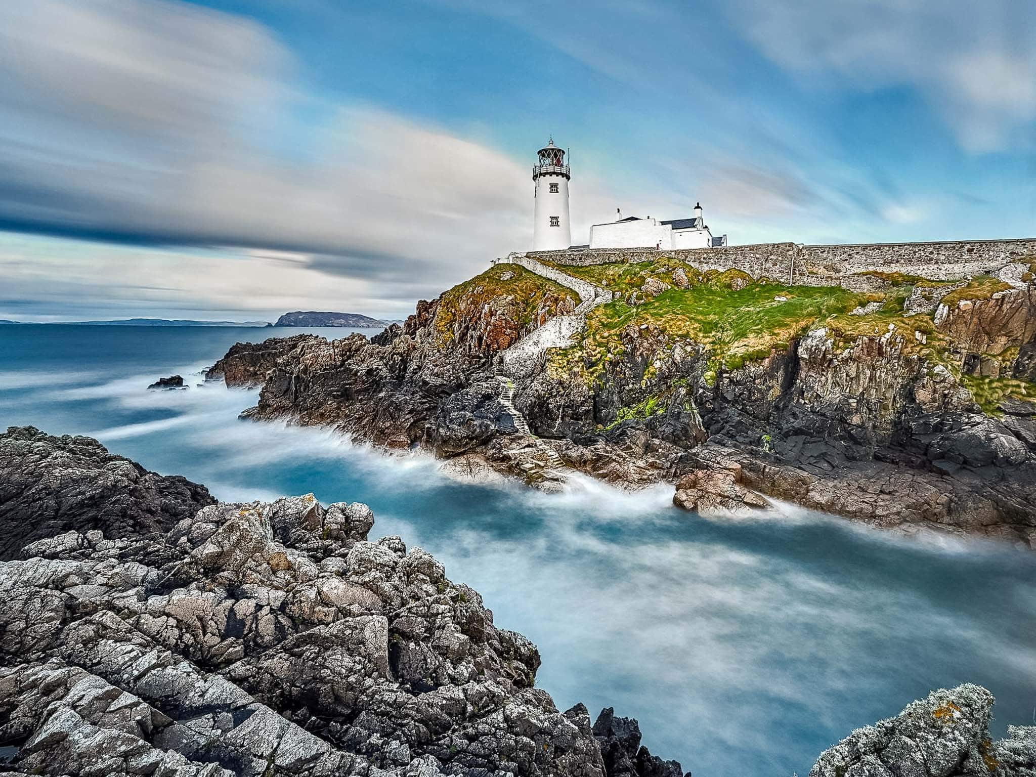 A white lighthouse stands on rocky cliffs against crashing waves and dramatic skies