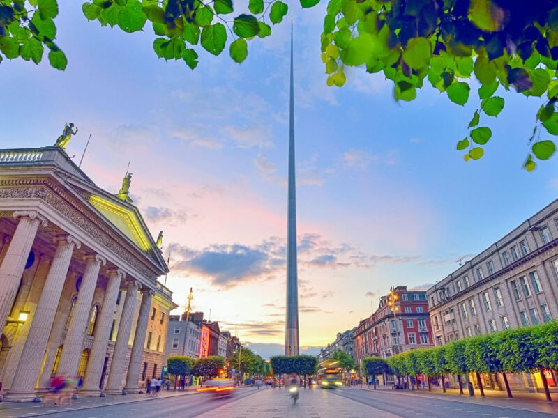 A wide street in Dublin with pastel coloured buildings lining each side and the Spire of Dublin rising dramatically into the evening sky in the background