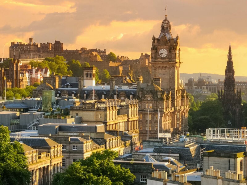 A panoramic view of Edinburgh over the rooftops with the Balmoral Hotel clock tower centre, Edinburgh Castle in the background, and the Scott Monument rises prominently to the right.