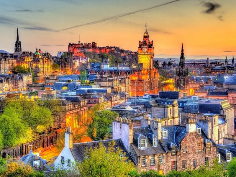 The skyline of Edinburgh glows at sunset, with the castle, church spires, and stone buildings lit by warm golden light.
