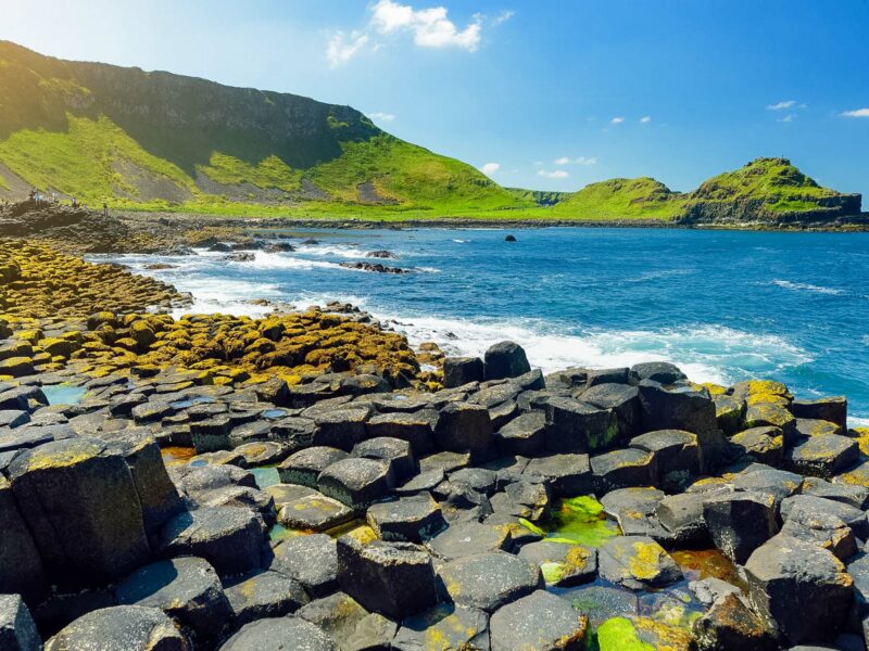 Basalt rock columns of the Giant’s Causeway stretching into the sea, with waves breaking against the rocks and grassy cliffs in the background.