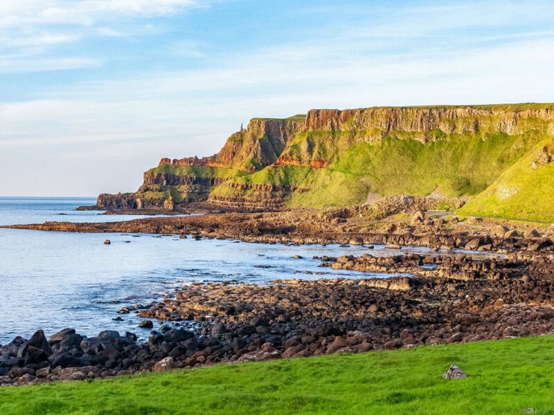 Coastal view of the Giant’s Causeway showing irregular basalt columns leading to the water, surrounded by green hills and a cloudy blue sky.