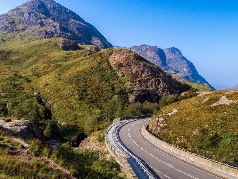 A curving mountain road threads through steep, grassy slopes in Glencoe, Scotland.