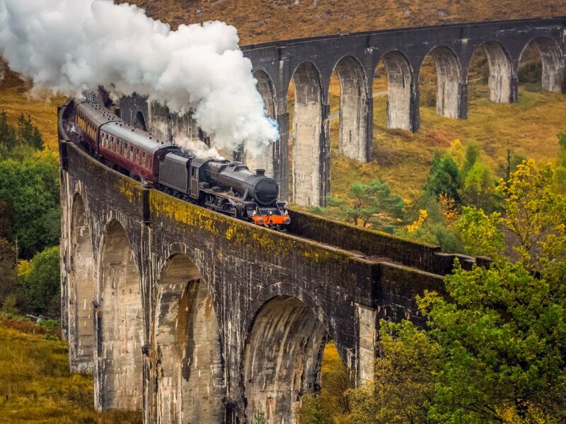 A black steam engine pulls carriages across the sweeping stone arches of the Glenfinnan Viaduct in Scotland
