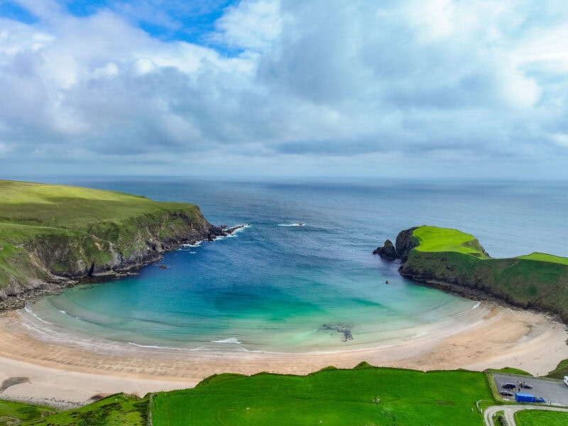 Towering green cliffs surround a bay of turquoise water with a sandy beach, leading out to the Atlantic Ocean extending to the horizon on a cloudy day.