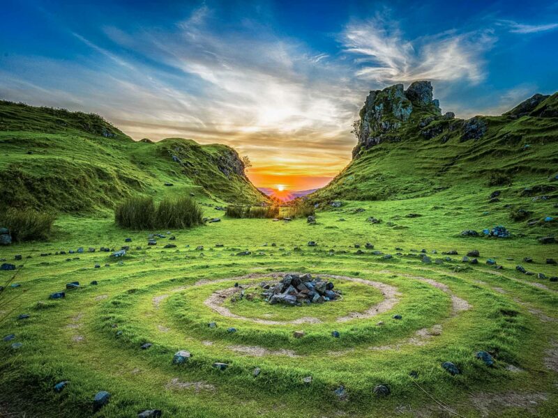 A circular stone arrangement on a grassy hillside, surrounded by lush valleys, under a golden sunset sky.
