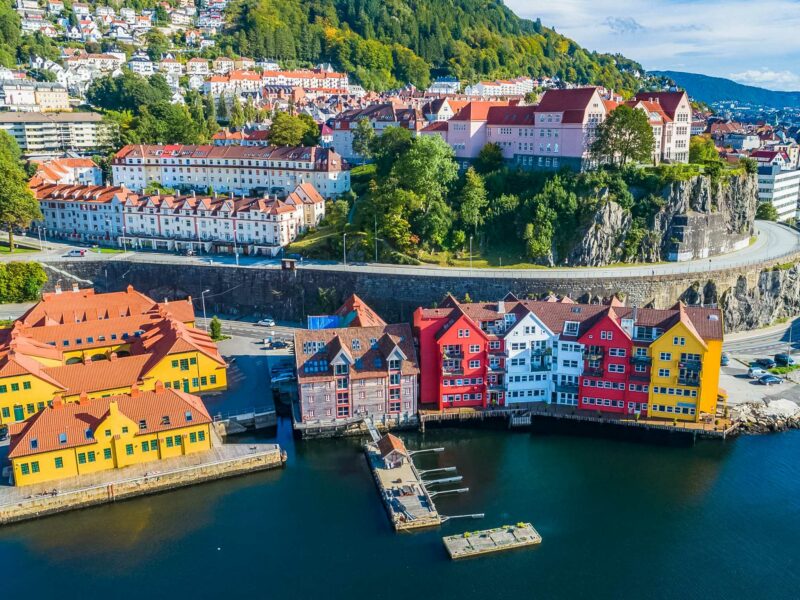 Colourful houses with red, yellow, and white facades lining the waterfront in Bergen, Norway, backed by hillside homes and lush green trees.
