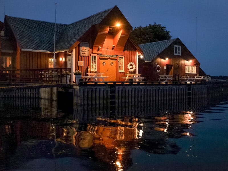 Red wooden buildings along the waterfront at night, warmly lit with reflections shimmering in the calm water, creating a cozy and inviting atmosphere.