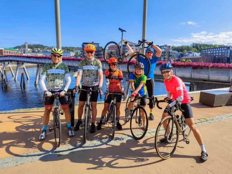 Six cyclists posing with bikes near a modern waterfront bridge in a Norwegian city, smiling under a clear blue sky. One rider holds their bike aloft.