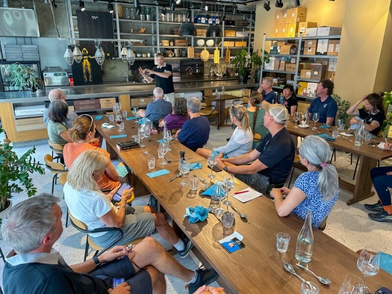 A group of people seated around wooden tables listening to a guide briefing in a modern kitchen with shelves, plants, and cooking equipment.
