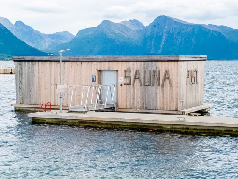 A wooden floating sauna house marked “SAUNA” moored on calm blue water, surrounded by towering mountains in the background.