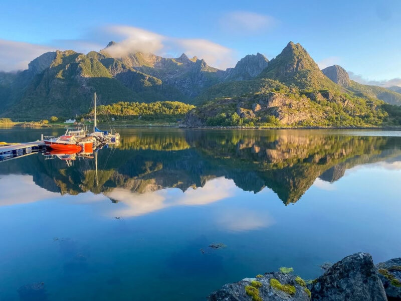 A glassy surfaced fjord with boats tied to a dock, the surrounding mountains and clouds reflected perfectly in the still water at golden hour.