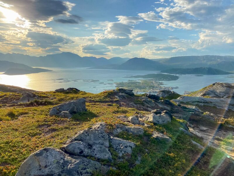 A dramatic sunset view from a rocky hillside overlooking fjords, islands, and shimmering water with golden light streaming through clouds.