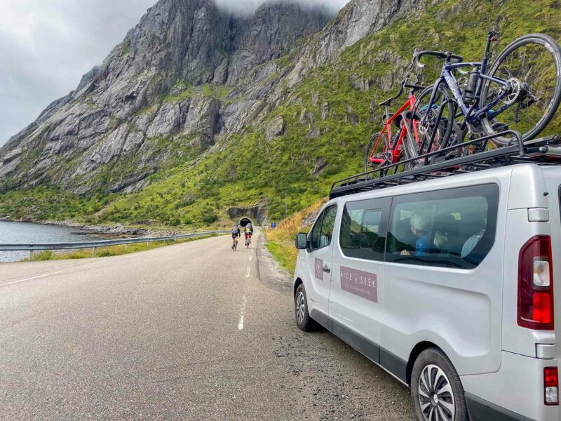 A support van with bikes on the roof parked by the roadside while two cyclists ride toward a tunnel carved into a steep rocky mountain.