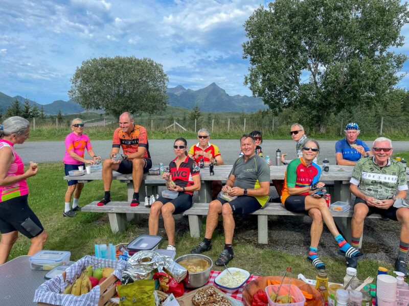A group of cyclists in colourful jerseys sitting smiling at a picnic table enjoying lunch, with food spread out on a second table in the foreground and mountains in the background.