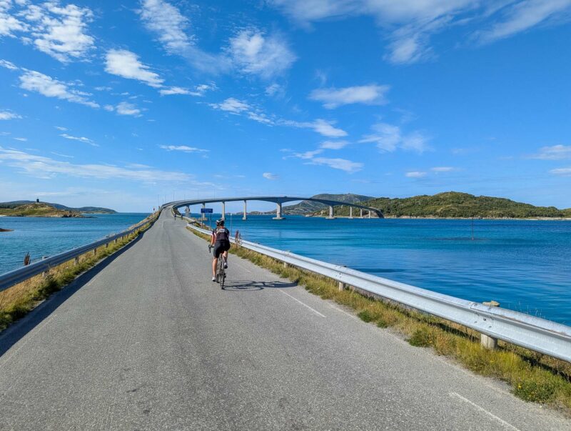 A lone cyclist rides along a causeway toward a long arched bridge over turquoise waters, with islands in the distance under a bright sky.