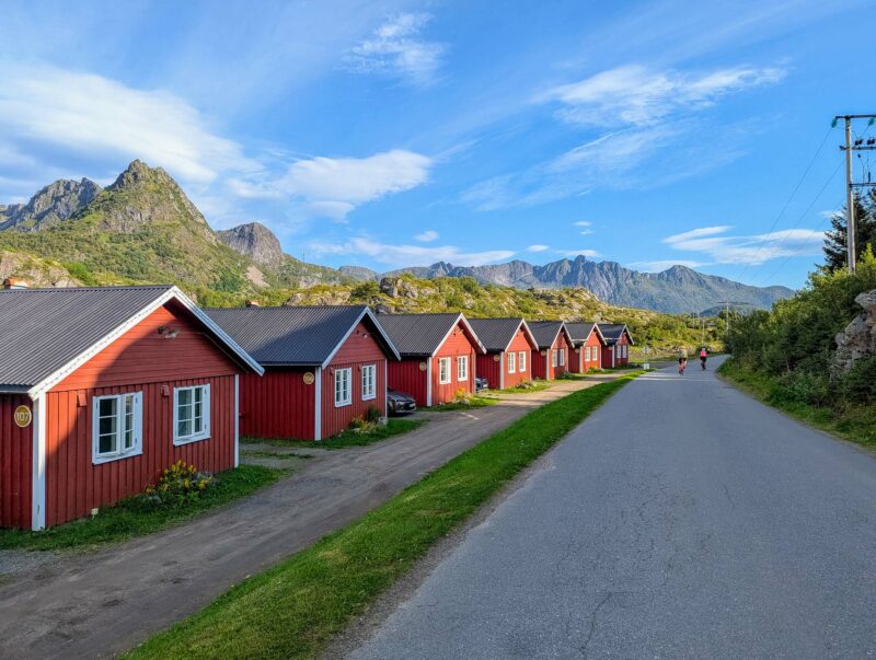 A row of red wooden cabins with white trim lining a quiet road, set against a mountain backdrop with green forests and clear skies.
