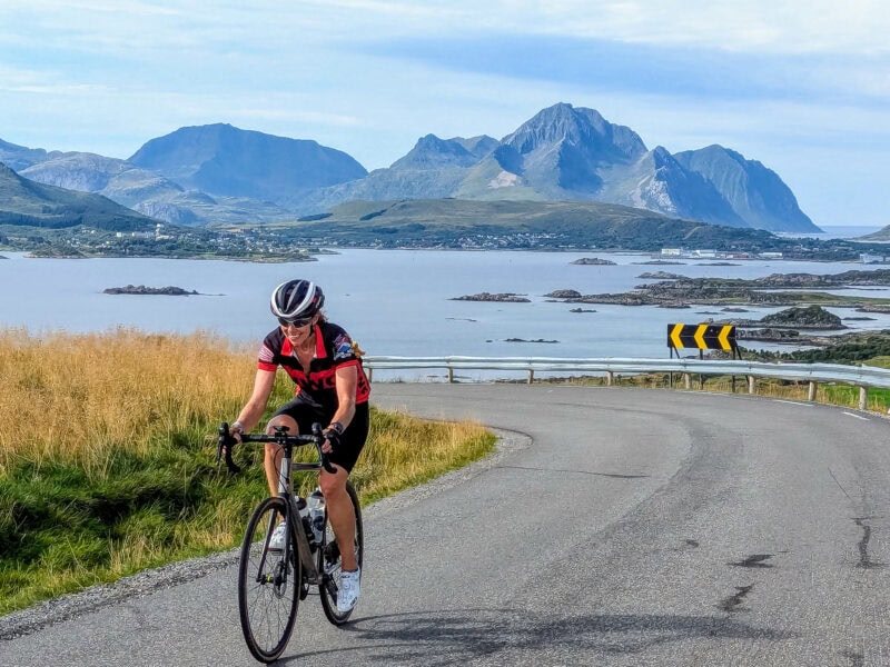 A cyclist in a red jersey climbs a winding coastal road overlooking calm waters and distant rocky mountains.