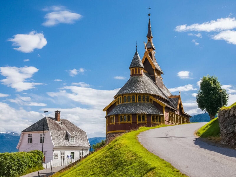 A traditional wooden stave church with ornate spires stands on a grassy hill under a blue sky.