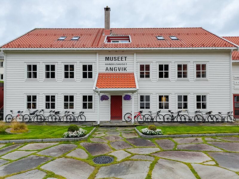 White wooden building with a red roof, the Angvik Handelssted Museum in Norway, with a row of bicycles lined up in front on a stone-paved courtyard.
