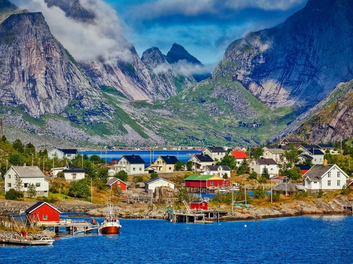 A picturesque Norwegian fishing village with colourful houses and boats by the shore, surrounded by dramatic mountains and low-hanging clouds.