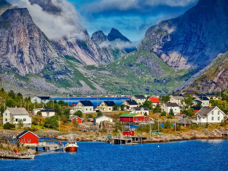 A picturesque Norwegian fishing village with colourful houses and boats by the shore, surrounded by dramatic mountains and low-hanging clouds.