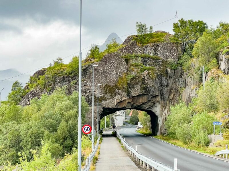 A mountain road passing through a natural rock tunnel, framed by greenery and cloudy skies.